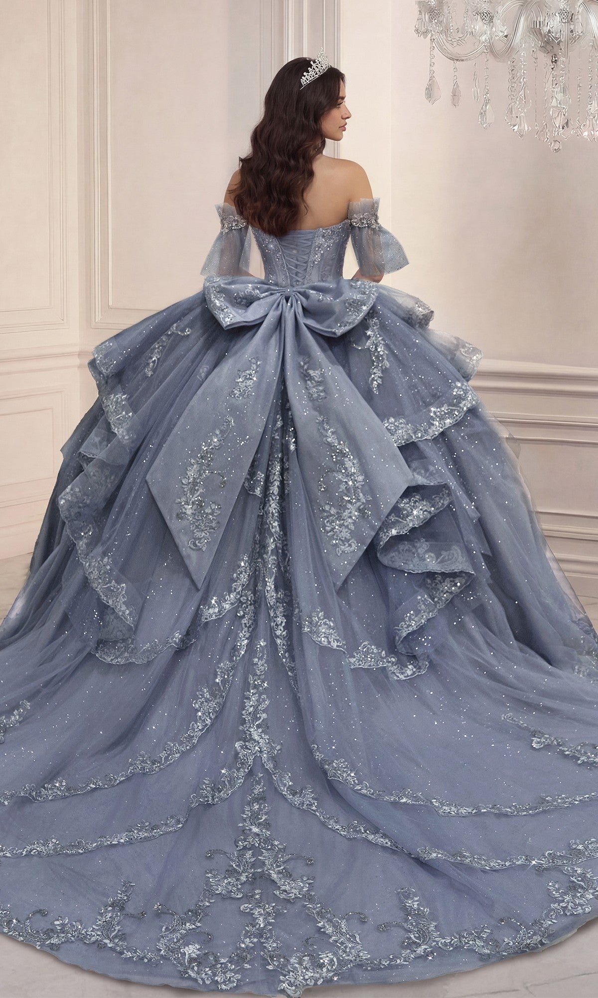 A woman wearing the Quinceanera Dress 15741 By Ladivine, a pale blue gown with lace, sequins, and glitter tulle, stands with her back to the camera in an elegant, light-colored room.